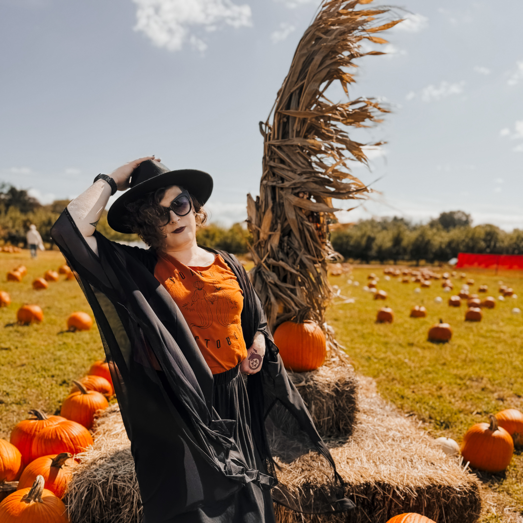 a photo of parker lee standing in a pick-your-own pumpkin patch. behind her is assorted pumpkins, bales of hay, and cornstalks. they are dressed in a black, wide brimmed hat, a black maxi skirt, an orange t-shirt, and a black mesh covering.